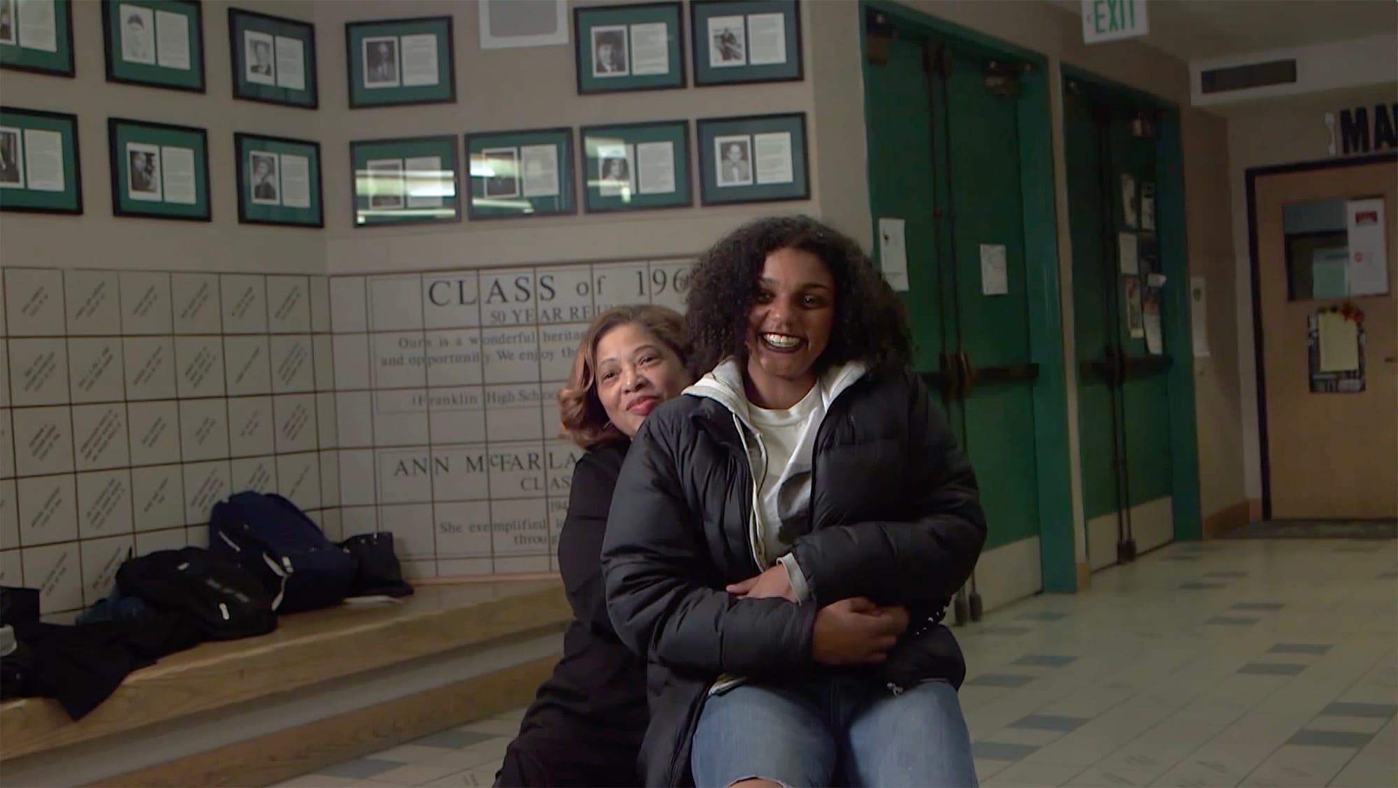 Woman, seated, with younger woman on her lap, in the hallway of a school.