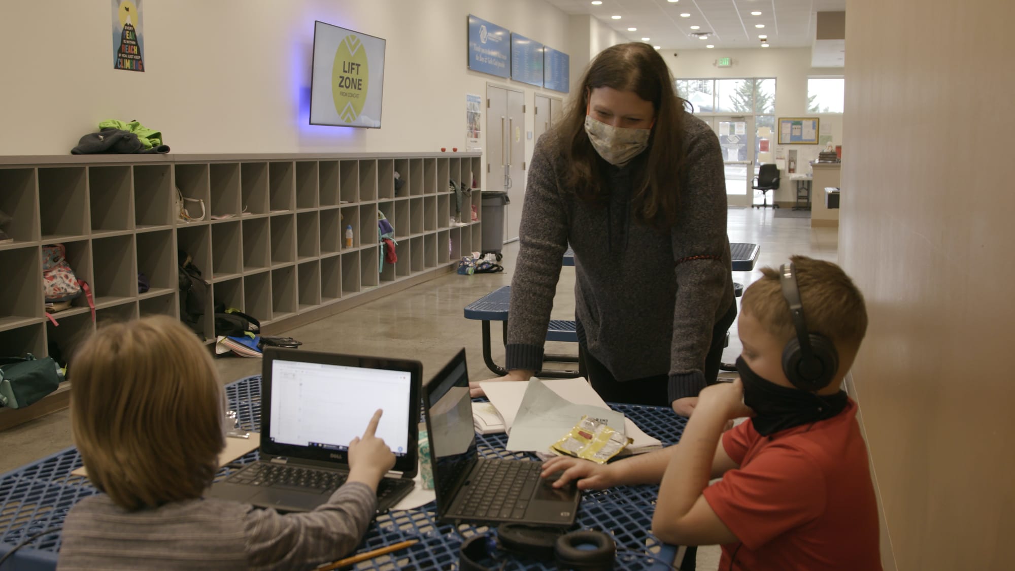 A teacher wearing a mask helps two students at a table