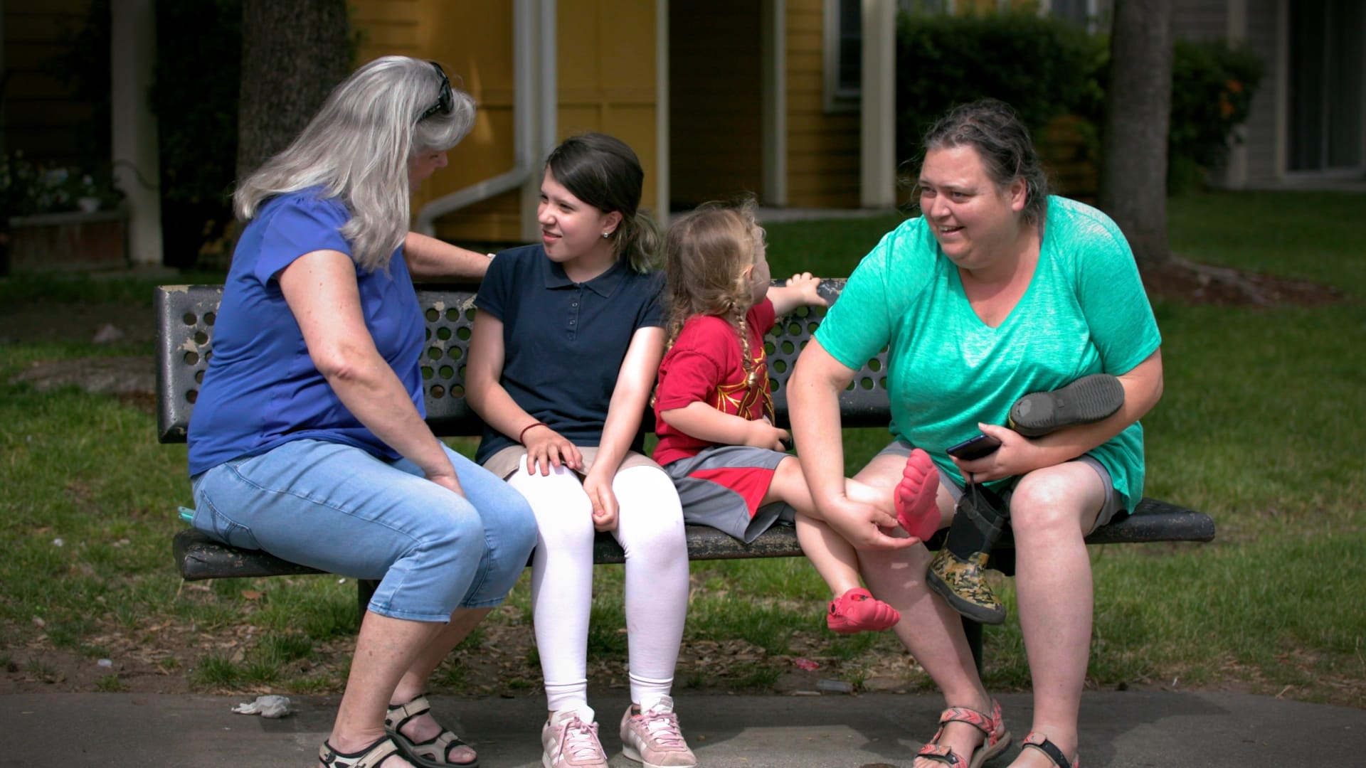 A grey haired woman, Julie Brenaman, sits on a bench talking with two children and their grandmother, who's trying to put a shoe on the smaller child.