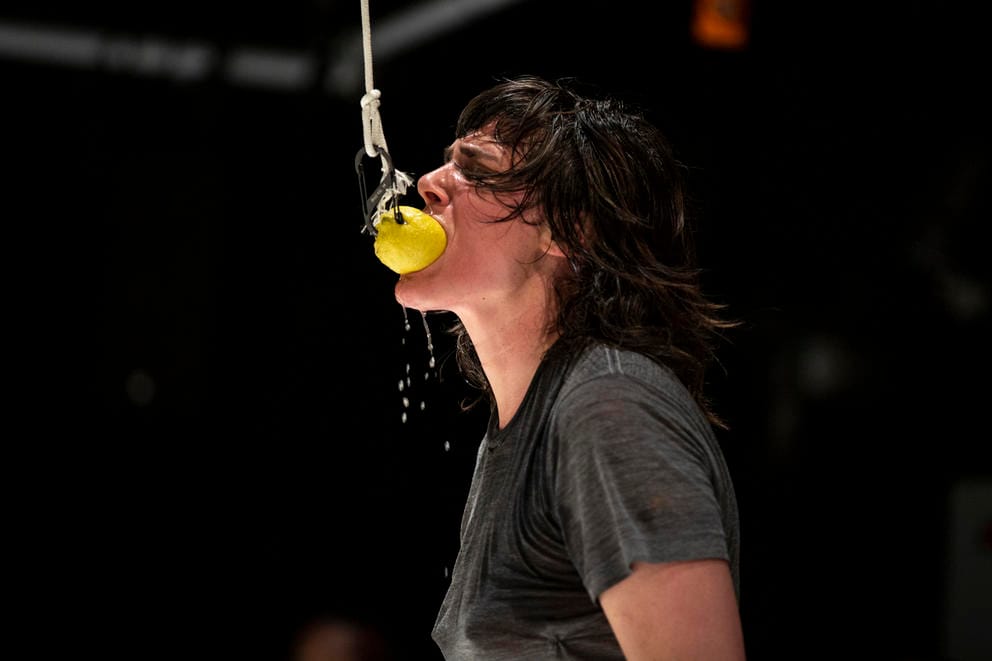 Person in gray t-shirt on black background bites in a lemon suspended from the ceiling