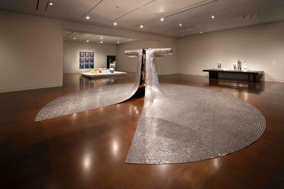 Museum gallery featuring, in the middle, a large sculpture that looks like a robe made from metal, its long sleeve extending over the floor