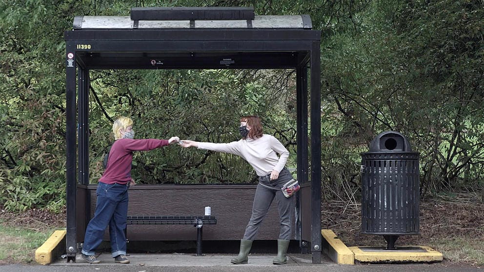 Two people in front of a bus stop, handing over hand sanitizer