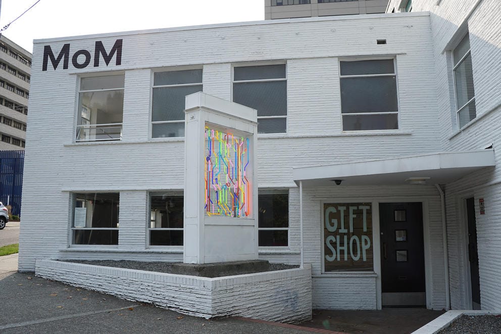 White building with a neon artwork out front. On the upper left corner, the white wall says MoM