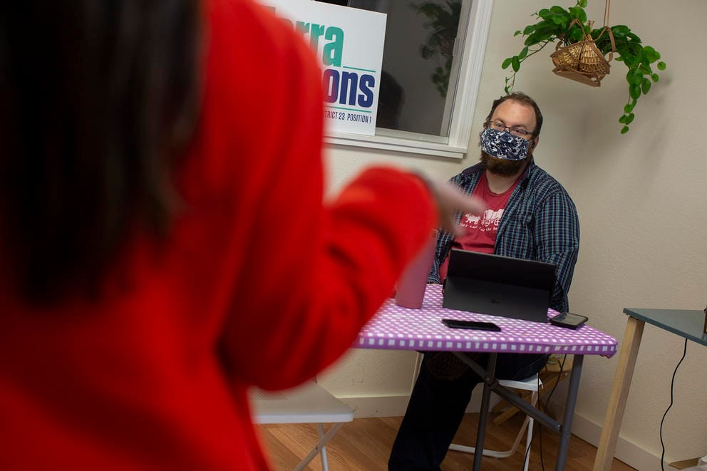 A woman with her back to the camera talks to a man seated in front of a laptop