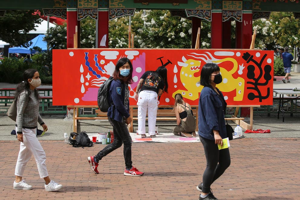 People walk in front of a large mural being painted on a wide wooden panel standing up