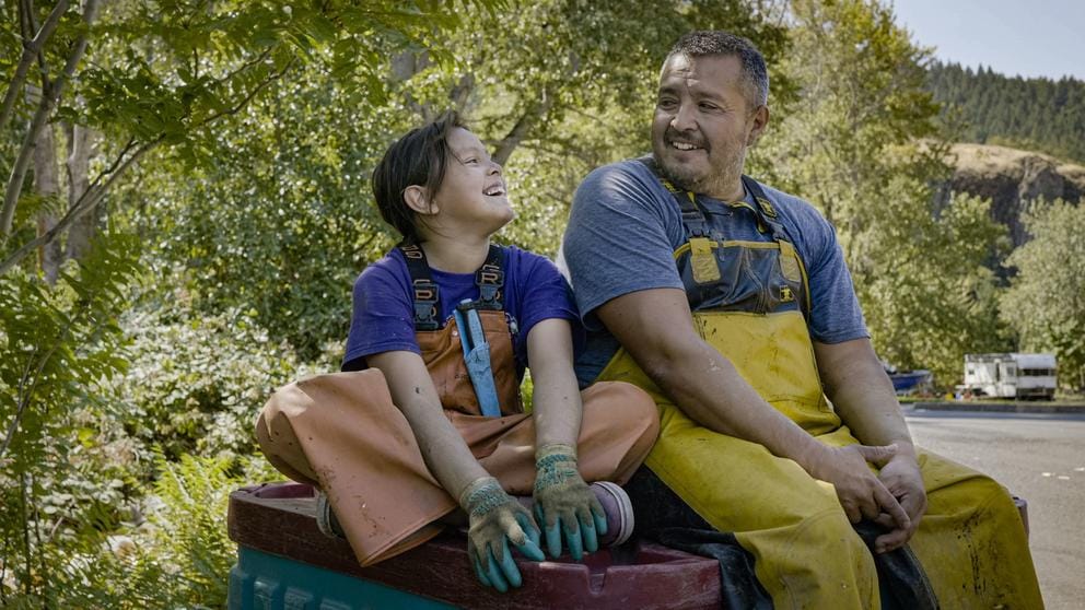A young girl sits cross-legged while smiling up at her father sitting next to her. Both wear fishing waders.