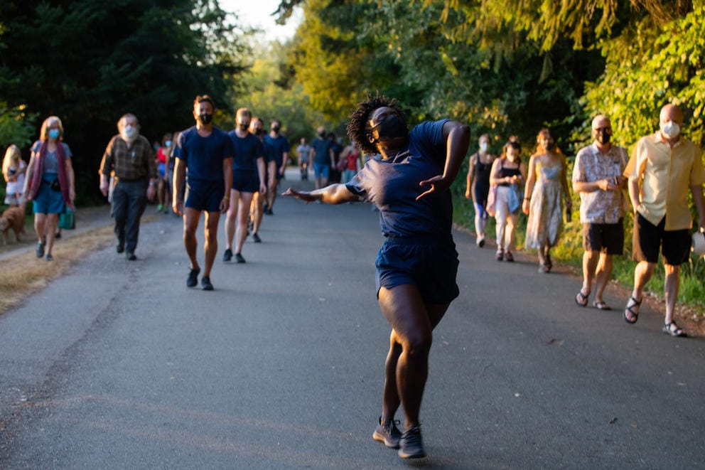 Person in dark blue outfit in middle of a park road dancing, people watching from the side