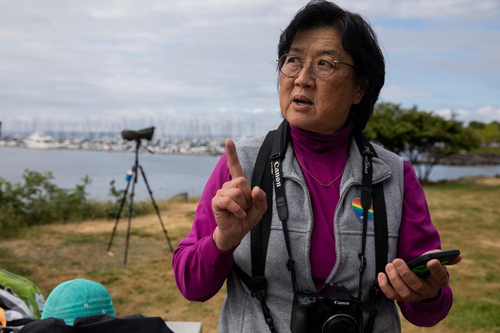 A person wearing binoculars standing in a park with a bay of water behind her