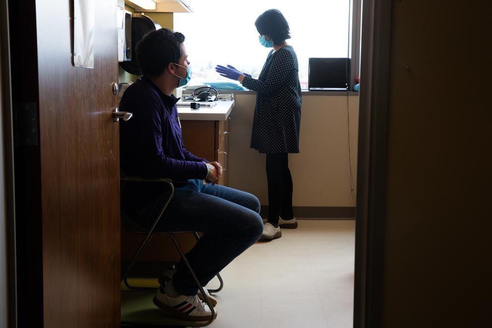 man sits in a chair in a doctor's office while a woman puts on gloves behind him