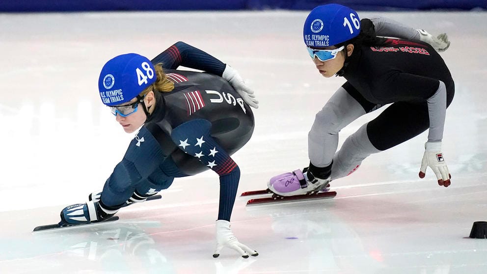 Two speedskaters in dark uniforms and blue helmets are on the ice, one following the other, both of their hands on on near the ice as they make a turn