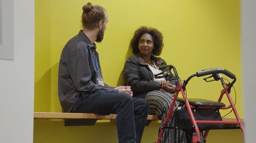 A man and woman sit on a bench, looking at each other, speaking