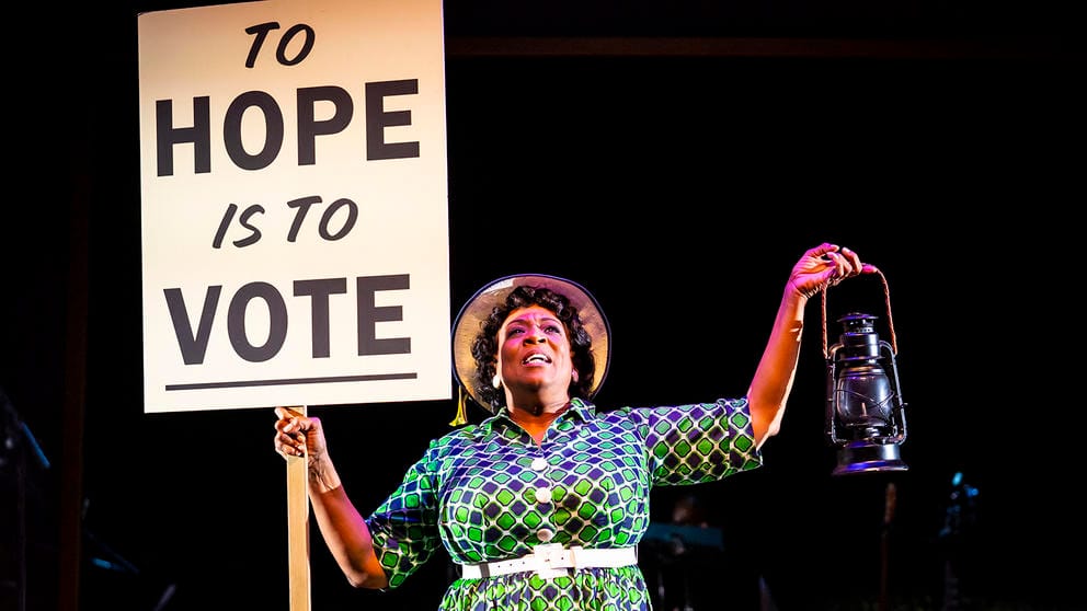 a woman in costume on stage holds a sign reading To Hope Is To Vote