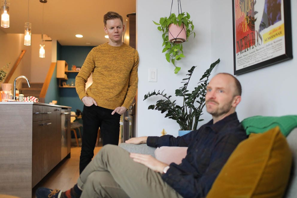 Ryan Hicks sits on a couch in the foreground as Jeff Dorion stands leaning against a wall in the background