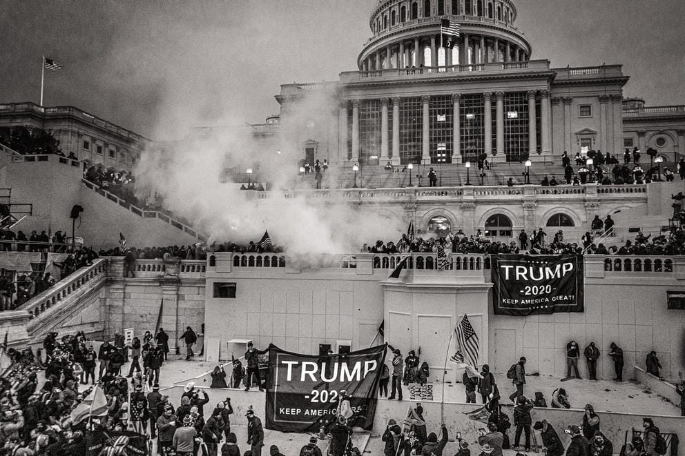 The capitol building is seen from afar, with the steps and platforms visible, there are people and flags and there is smoke from teargass, in a black and white photo