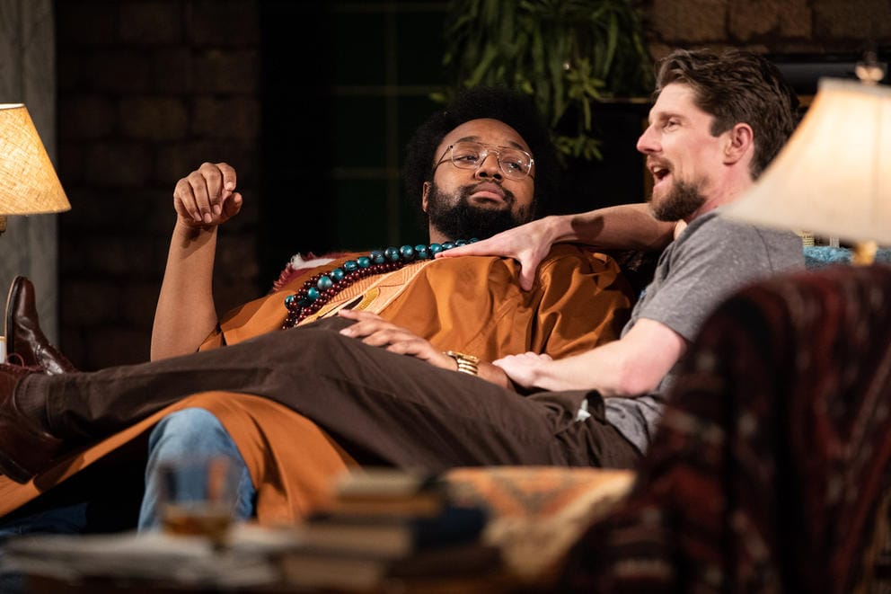 A man in an orange shirt lies back on a couch while a man next to him puts his legs over his lap