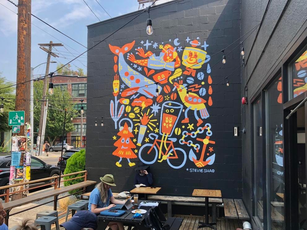 Cafe with person sitting behind laptop, outside, in front of a tall black wall featuring a colorful mural with animals and bikes.