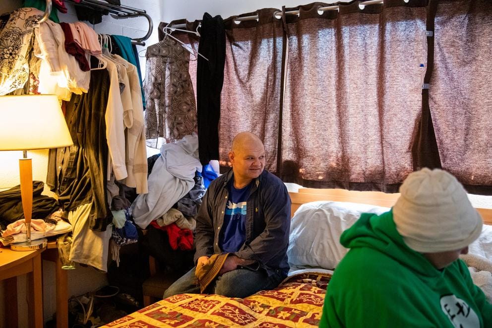 Man sits on a bed watching TV that is not in the picture, looking over his left shoulder. A woman sits on the other side of the bed looking at the TV that is to the right of the picture frame. Clothes hang in the background at left