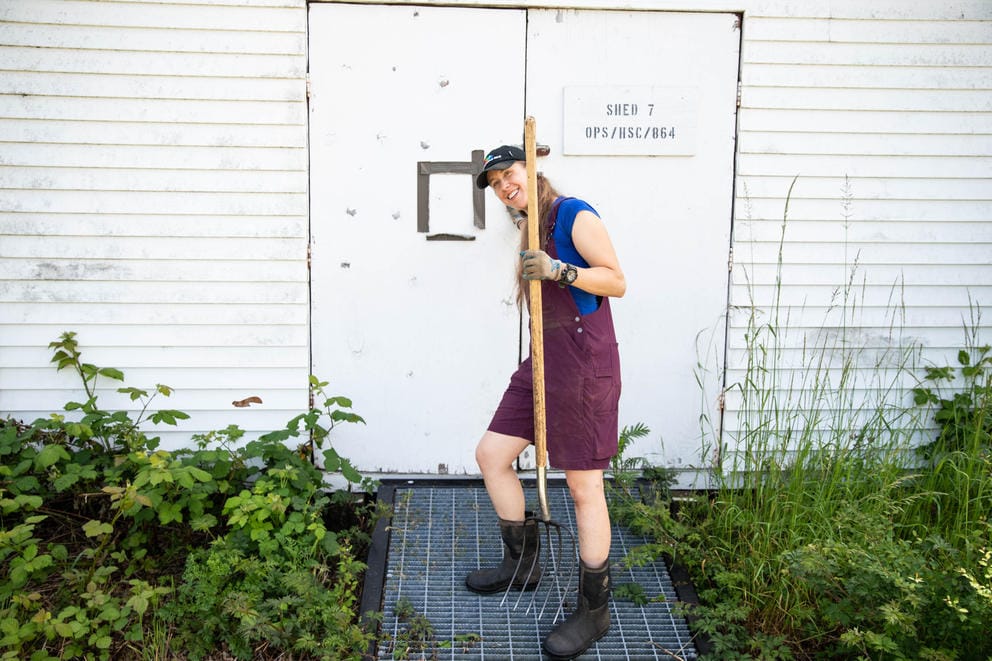 A person in purple overalls with long hair closes the door to a white shed while holding a shovel.