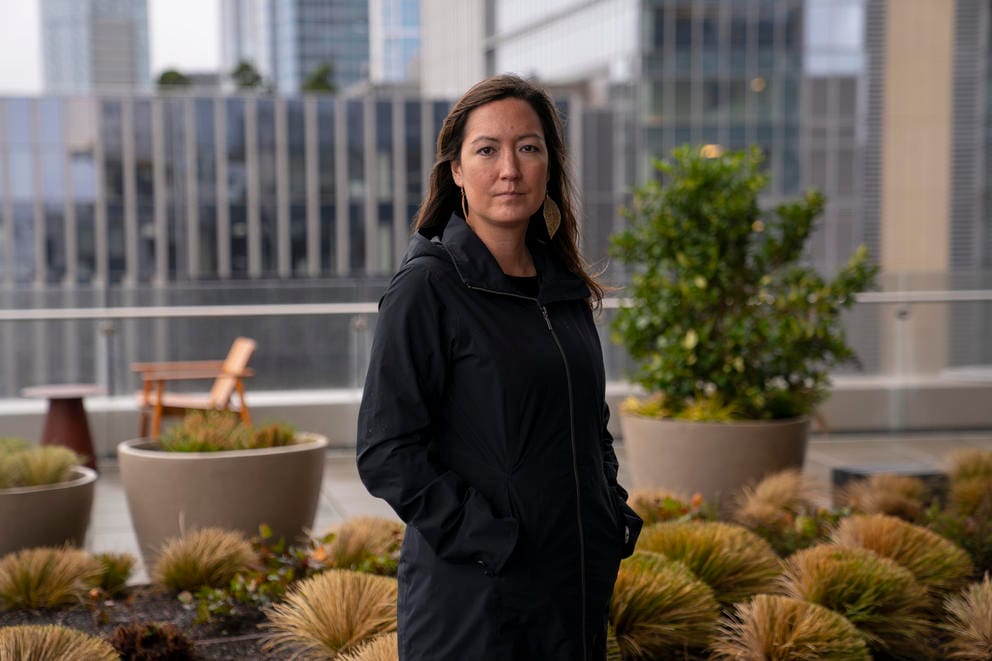 A woman in a black coat stands with her hands in her pockets on a rooftop