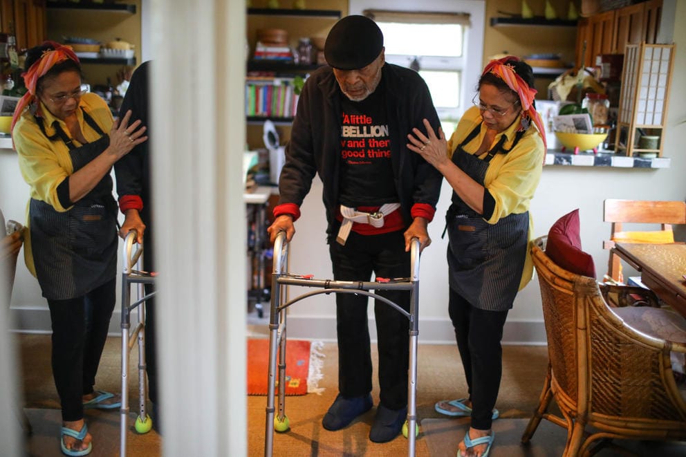 A woman helps a man walk with a walker, their reflection caught in a large mirror