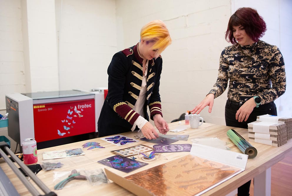 Two people standing at a large table, with a printer in the background. They are holding paper butterflies.