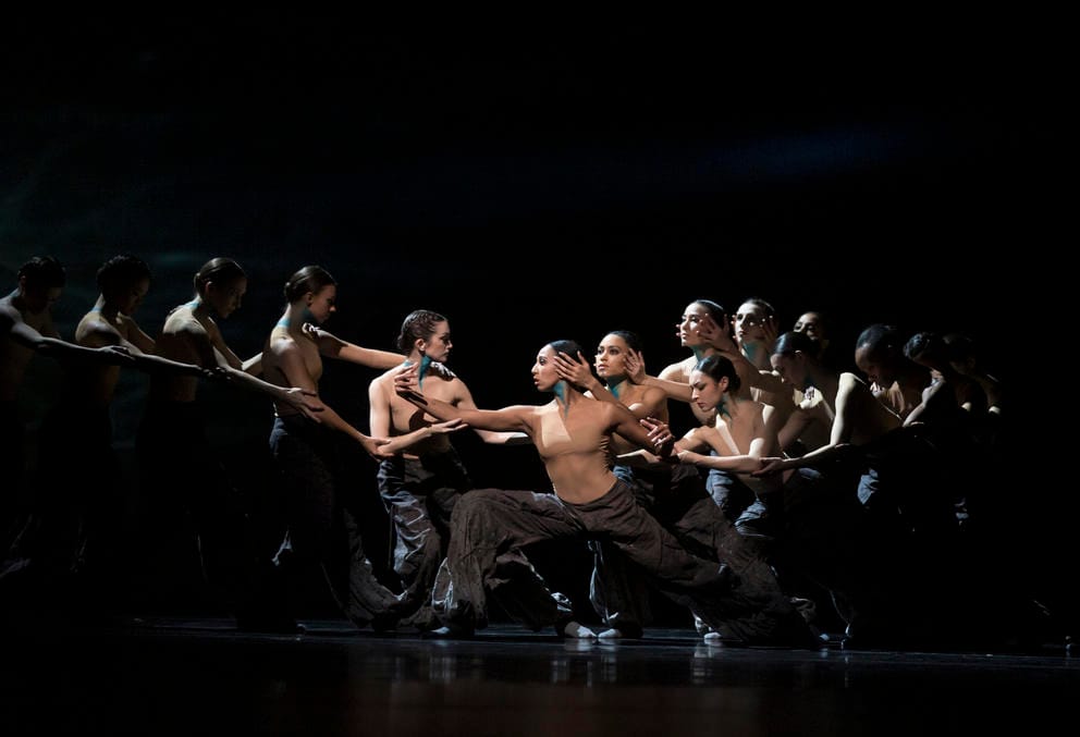 photo of dancers on a dark stage in two lines both facing a central dancer