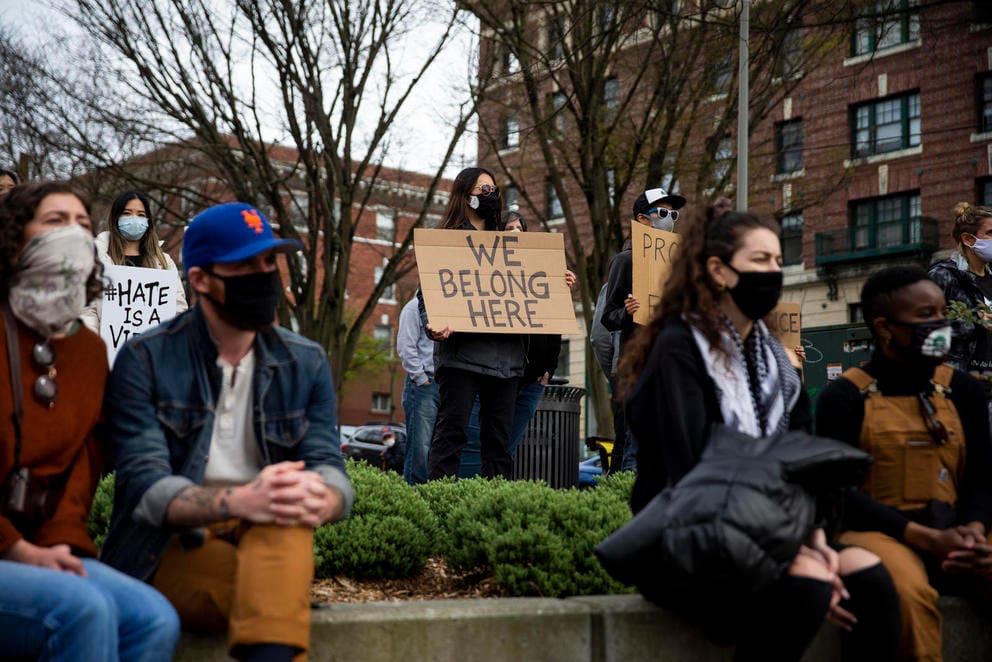 masked people gather at an outdoor rally, a central figure holds a sign that reads