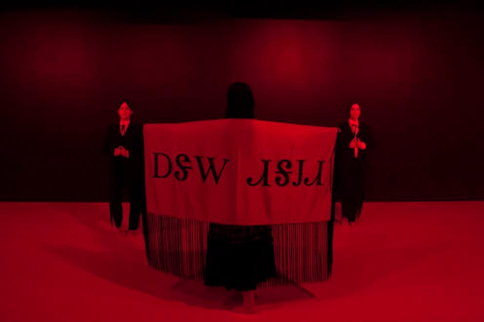 dark stage bathing in red light, one person is wearing with arms wide open a large piece of cloth. Two people in suits stand on either side, a little towards the back.