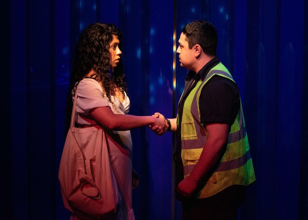 Two people on a stage in front of plastic blue backdrop. She is wearing a white dress and has long, black curly hair. He, shaking her hand, is wearing a yellow hi-viz vest.