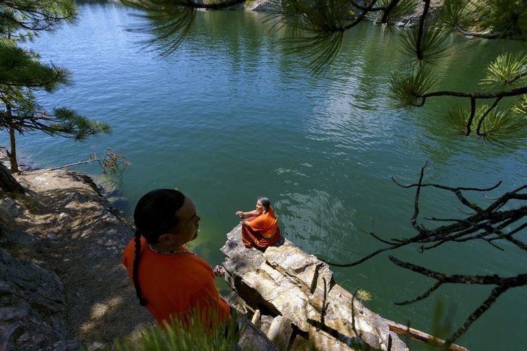 A woman sits on a rock on the water. A man stands on a ledge above