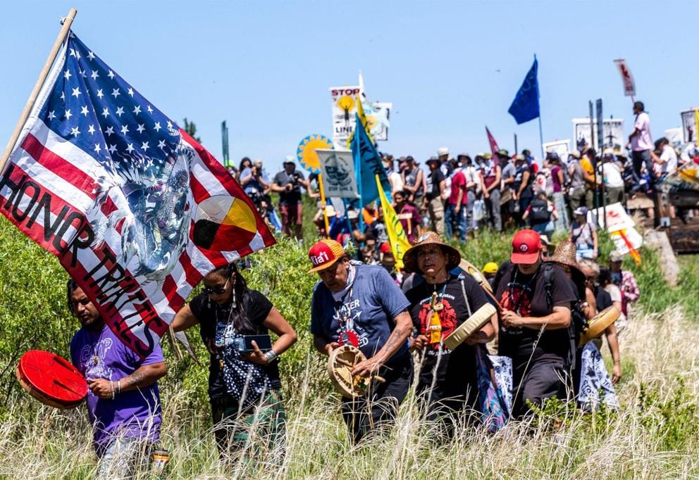 A group of Indigenous activists walk through tall grass holding flags and signs, one of which reads 