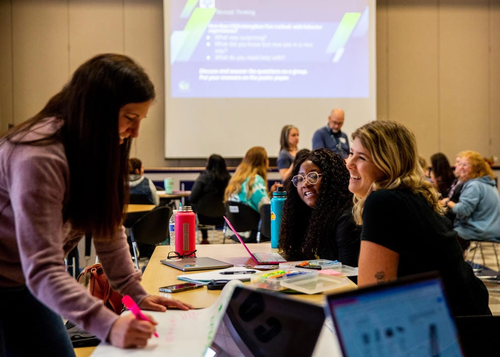 School employees work in small groups at a table covered in laptops, papers and markers.