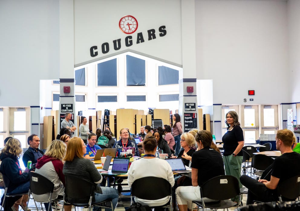 About a dozen school employees sit around a circular table with laptops as the word