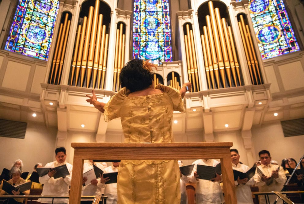 A conductor is seen from behind with arms raised, conducting a choir, with large organ pipes rising above them