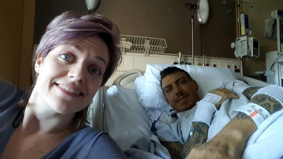 A woman smiles for a selfie with a man weakly smiling from a hospital bed.