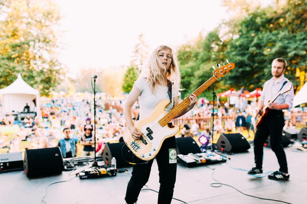 Person with blond hair, white tshirt and dark jeans looks at the camera, playing a guitar. A crowd is in the background. People are outdoors.