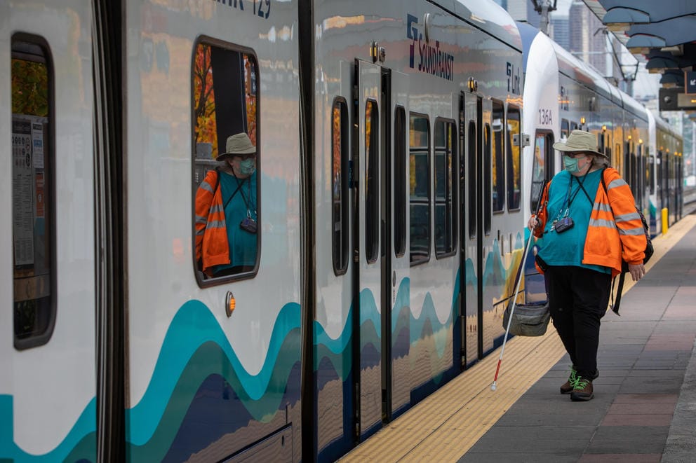 A wide shot of Dorene Cornwell in neon orange sweatshirt waiting at the Sodo Station platform as a train pulls in.