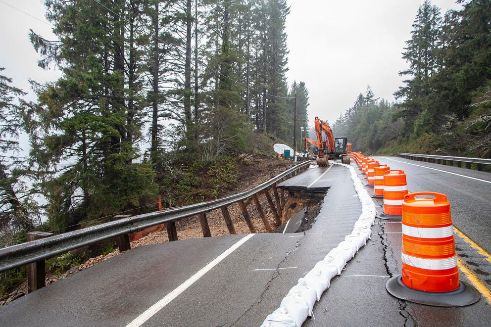 Large orange pilons line a highway where a chunk of the road has fallen down an eroding hillside