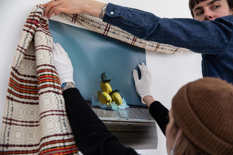 Two people work on hanging up an artwork, one is wearing white gloves and holds up a printed photo. The other person is holding up an embroidered red and white piece of fabric.