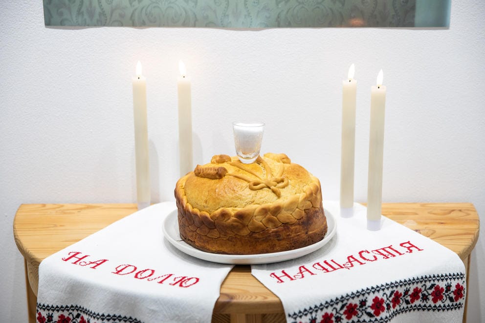 A round loaf of bread sits on a piece of fabric on a table, flanked by two candles on each side. On the fabric, red letters spell out: “na dolju” and “na shastja.