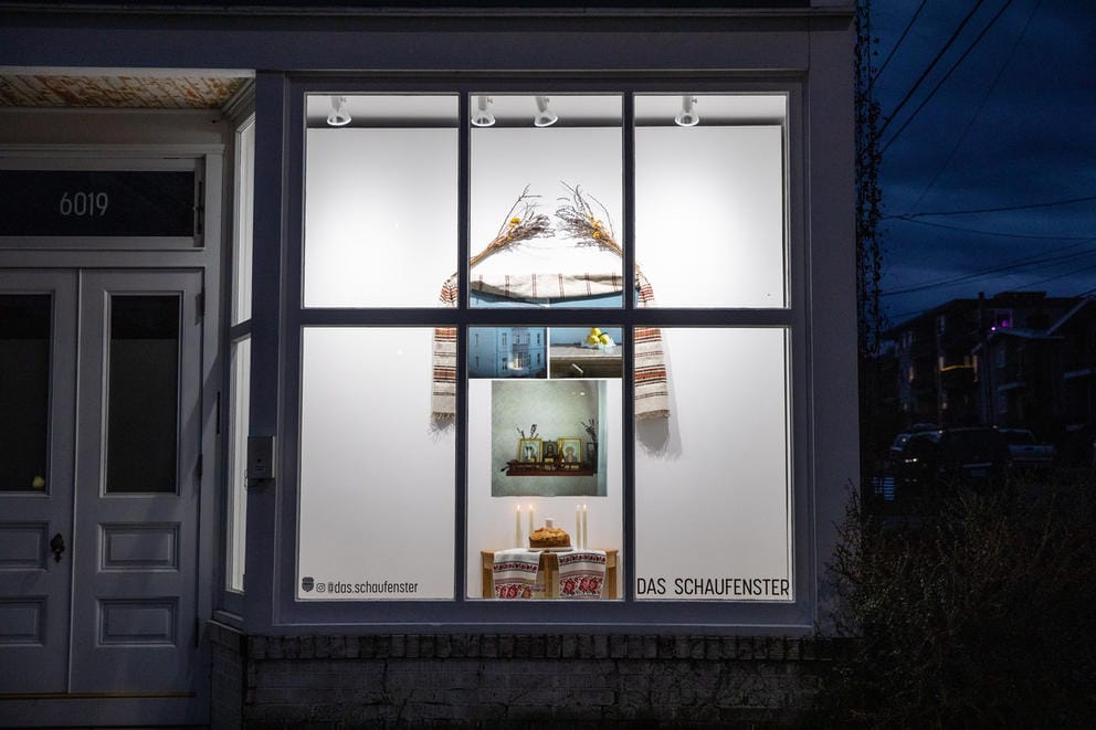 Window display seen from outside, where it is dark. Inside, three photos are framed by dried flowers and a piece of embroidered cloth.