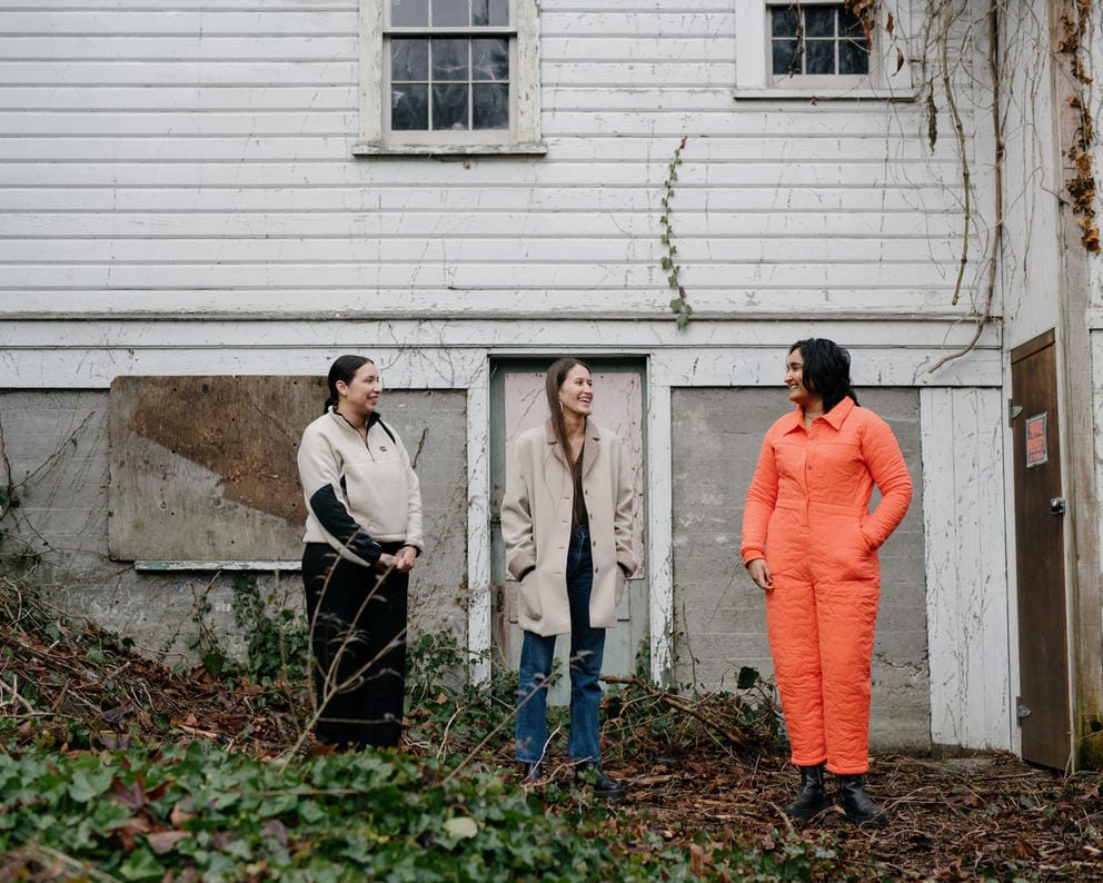 Three people stand in front of a shed, one person wears a beige fleece, the other a beige vest and the third person an orange jumpsuit