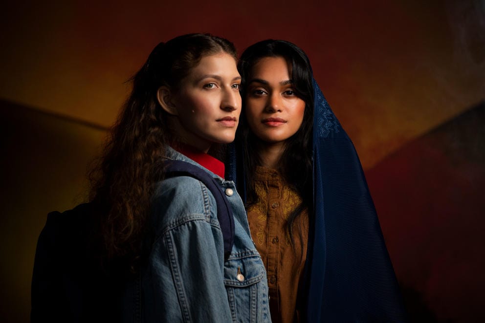 Two women stand next to each other, one looking at something beyond the camera and one making direct eye contact with the viewer