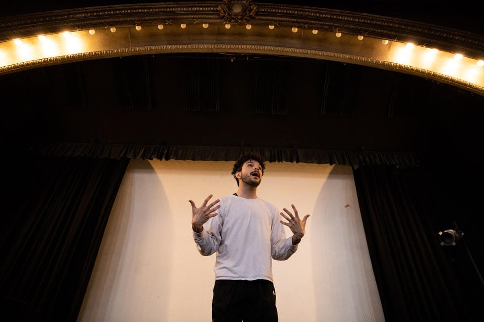 a man stands on a stage in a white t-shirt and black paints, talking