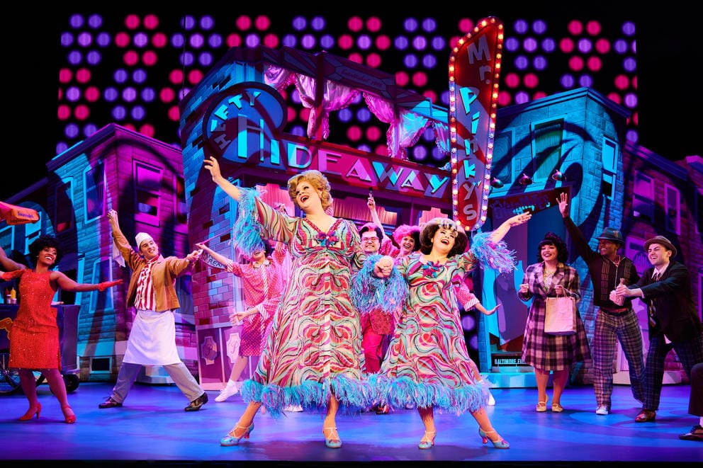 Two women in matching patterned dresses hold hands up and raise the opposite hands upwards on a wildly colored stage