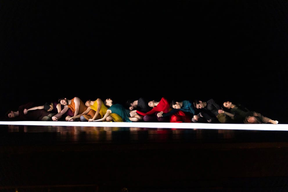 a dark stage in which a row of dancers in rainbow colored costumes sit at the edge of a ray of light