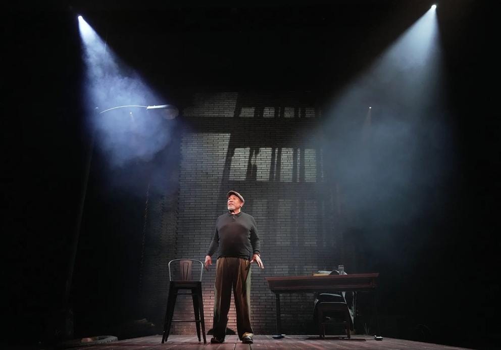 On stage, a man stands between a chair and a desk, facing the audience on a dark set