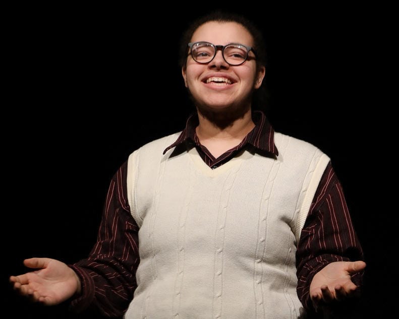 A boy in glasses, a white vest and dark shirt stands onstage with his hands gesturing outwards