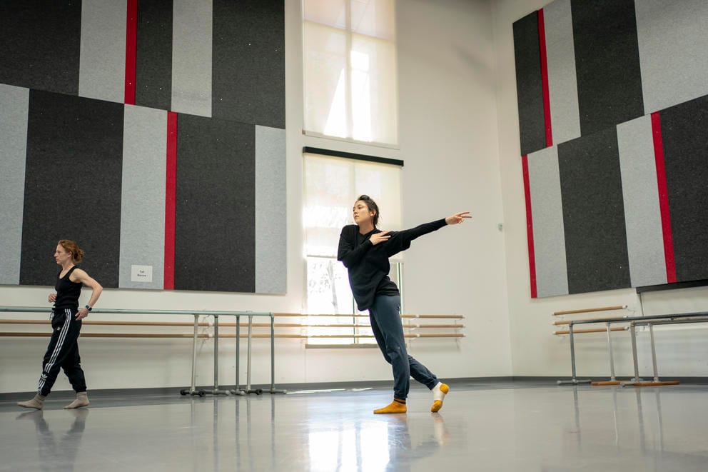A wide shot of a dancer rehearsing in a studio, the choreographer walks out of the frame at the left
