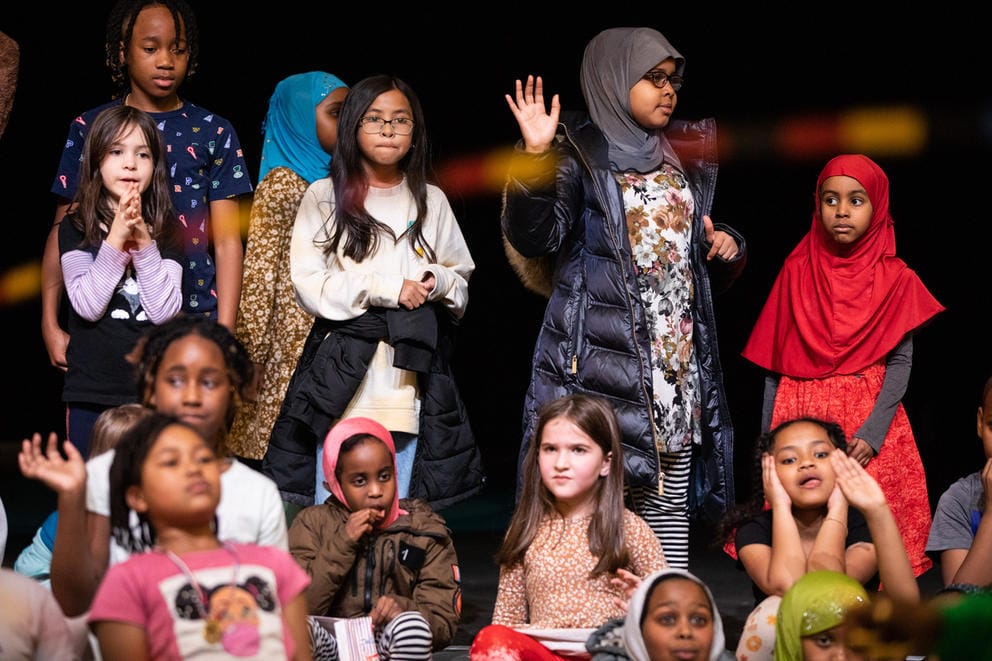 Twelve children stand or sit against a wall and listen intently to an off-camera speaker.